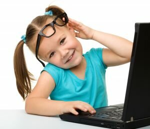 Cute little girl is sitting at table with her black laptop and wearing glasses, isolated over white